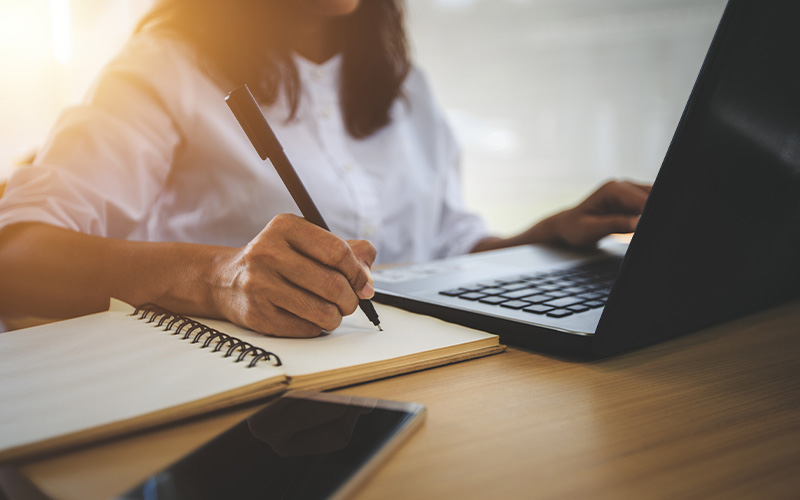 person writing in a notebook while using a laptop in a bright workspace focusing on productivity and organization for achieving goals in three steps